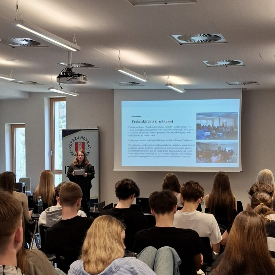 Skolniece prezentē savu darbu Konferencē, pārējie klausās, fonā ir prezentācijas slaids