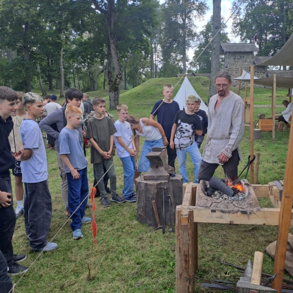 Skolēnu grupa, kas stāv ap vīrieti, kurš stāsta par kalšanas darbiem, fonā uguns