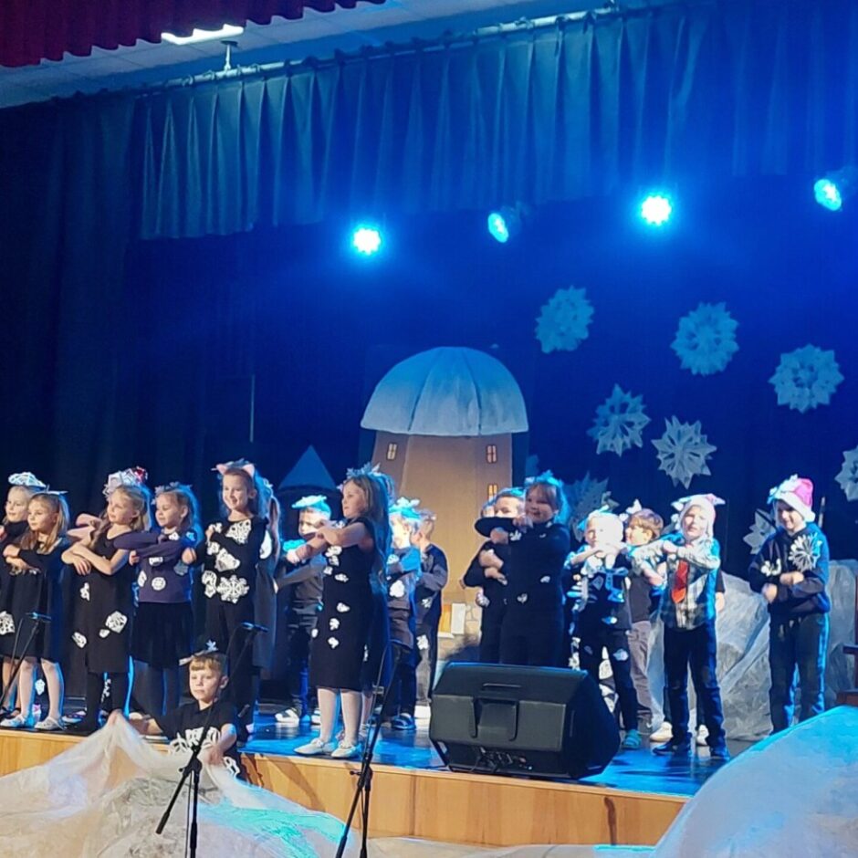 Bērni uz skatuves dzied, tērpi izrotāti ar sniegpārsliņām