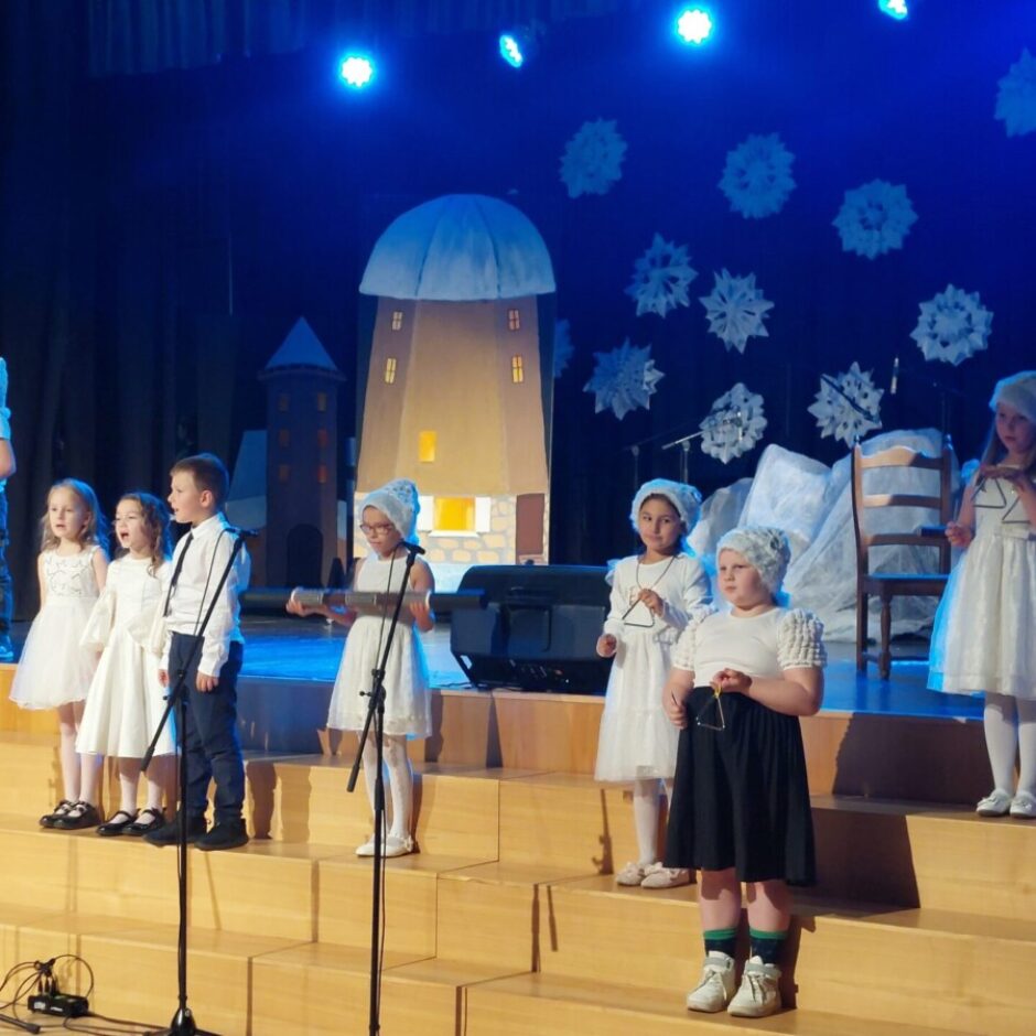 Bērni dzied uz Ziemassvētku skatuves, meitenes ir baltajās kleitiņās