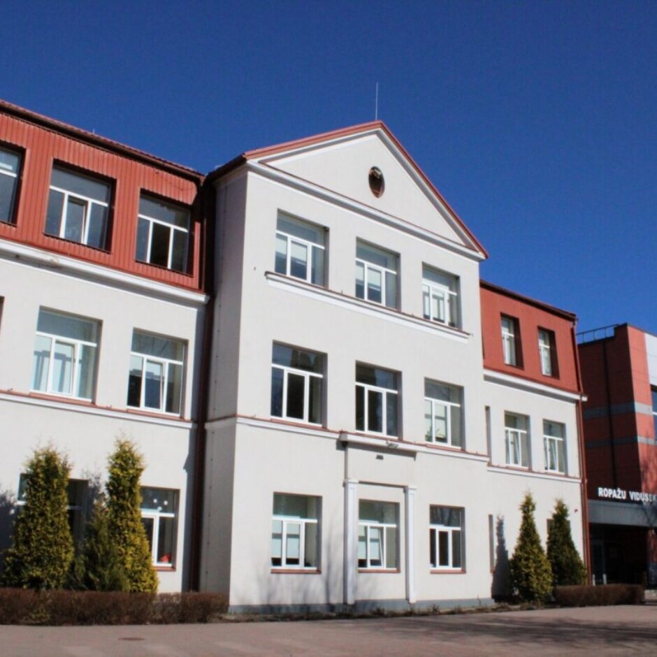 Ropažu vidusskolas ēka ar gaišu fasādi un sarkanbrūnu jumtu, pie kuras pievienota modernāka, sarkanbrūnas krāsas piebūve ar lieliem logiem. Pie ieejas uz fasādes lasāms uzraksts ROPAŽU VIDUSSKOLA. Fonā skaidras zilas debesis.