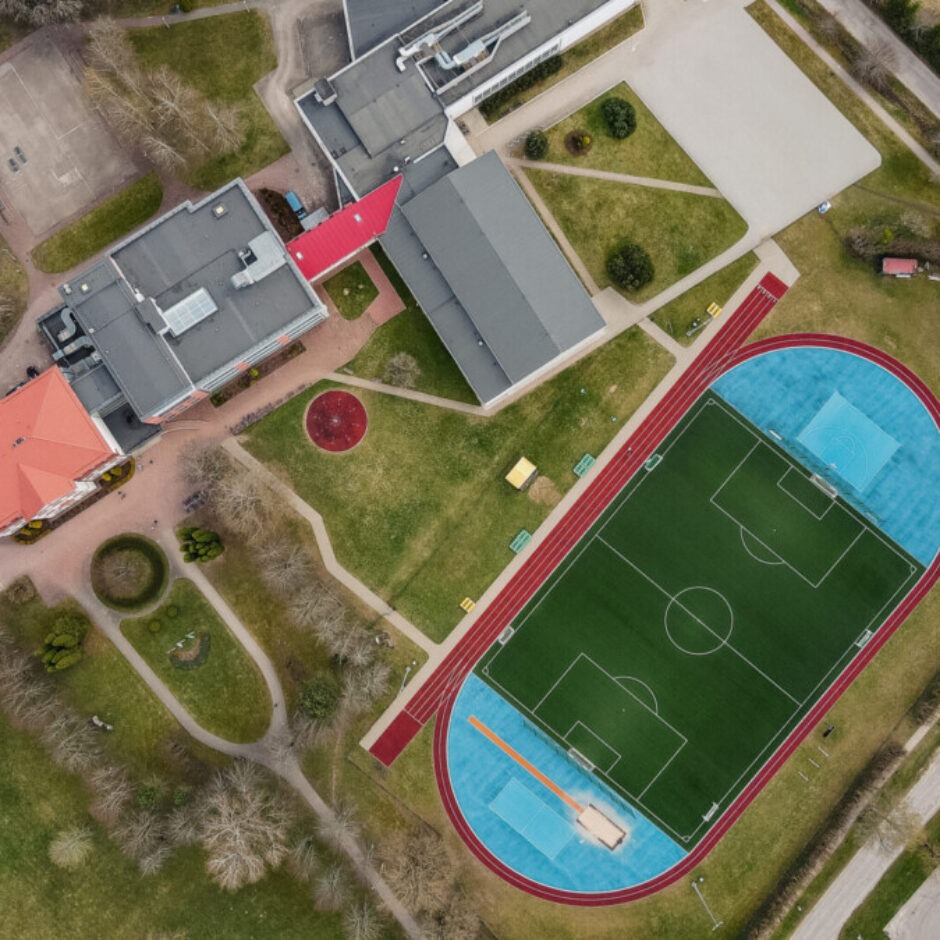 Aerofotogrāfija ar skolas vai sporta kompleksu, kurā centrā ir zaļš futbola laukums ar sarkanu skrejceliņu un gaiši zilu lēcienu zonu. Apkārt redzami vairāki ēku bloki, tenisa un basketbola korti, plaši zālāji, pastaigu takas, ceļi un autostāvvieta.