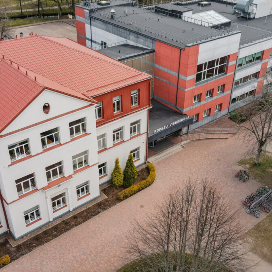 Skaists skats no augšas uz Ropažu Vidusskolu ar divām galvenajām ēkām – vecāku, baltu ar sarkanu jumtu un jaunāku, modernu ēku ar sarkanpelēku fasādi. Pie jaunākās ēkas ieejas redzams uzraksts 