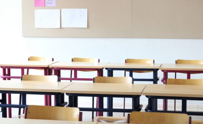 Klases telpa ar galdiem un krēsliem, fonā ziņojuma dēlis