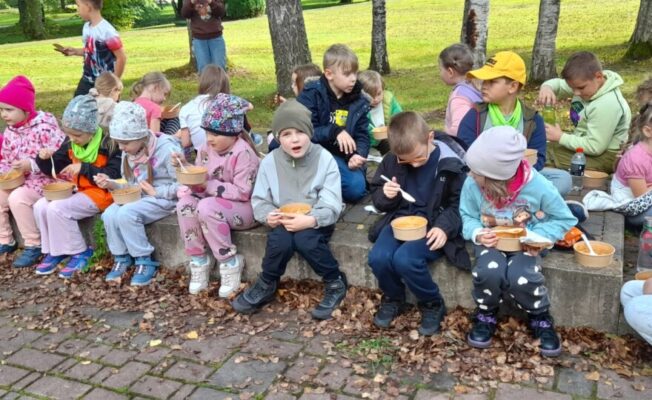 Skolēni sēž uz laukuma apmales un ēd zupu pēc pārgājiena.