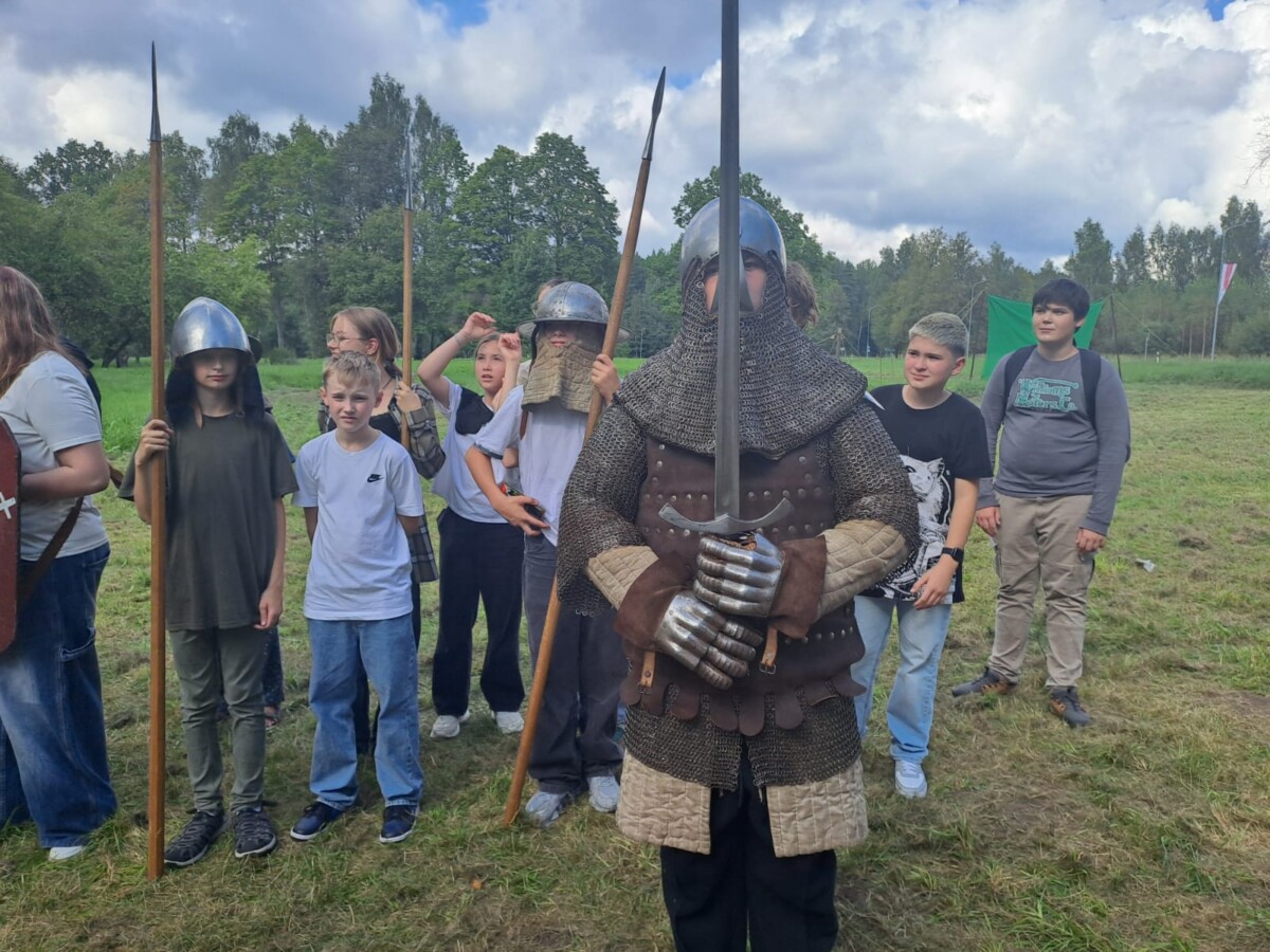Skolēni ar cilvēku, kurš pārģērbies par viduslaiku bruņinieku, kuram rokās ir šķēps