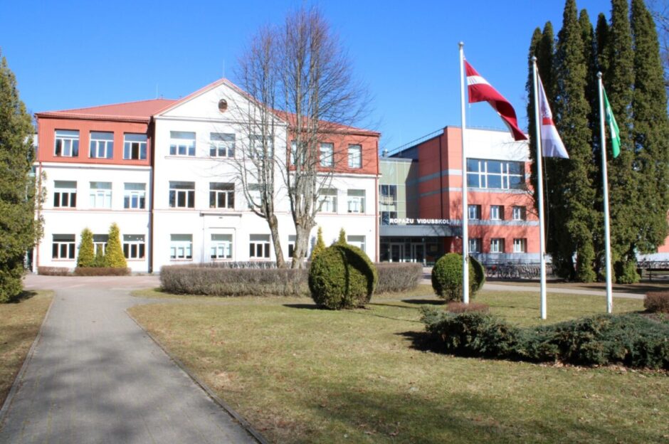 Daudzstāvu skolas ēka ar baltām sienām un sarkanu jumtu. Virs ieejas uzraksts 