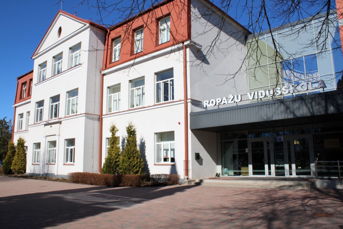 Ropažu vidusskolas ēka ar baltu fasādi un sarkanu jumtu. Virs galvenās ieejas ar stikla durvīm ir uzraksts 