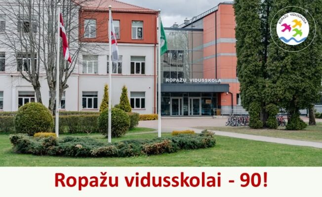 Ropažu vidusskolas ēka ar karogiem un skolas nosaukumu pie ieejas. Uz attēla redzams paziņojums: Ropažu vidusskolai - 90!